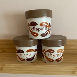 May include: Three ceramic spice jars with brown lids and white bodies. The jars have brown and orange leaf designs and the words "allspice", "sage", and "thyme" written on them.