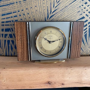May include: A vintage quartz clock with a gold-tone face and hands, set in a rectangular frame. The clock face has Roman numerals. The frame has a silver centre and wood-grain side panels. The clock is labelled "ME TAMEI QUARTZ".