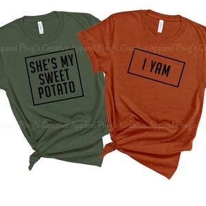 May include: Two short-sleeved t-shirts, one olive green and one burnt orange. The olive green shirt has the text "SHE'S MY SWEET POTATO" in a black box. The orange shirt has the text "I YAM" in a black box. Both shirts are knotted at the bottom.