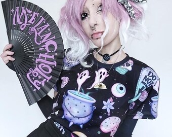 May include: A black fan with the word "Melancholy" in purple script is held in front of a person. The person is wearing a black top with a colorful print of ghosts, cauldrons, and other spooky elements. They are also wearing a black skirt.