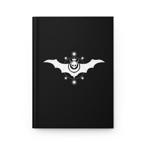 May include: Black notebook with a white bat and skull design. The bat has a crown on its head and is surrounded by a circle of stars.