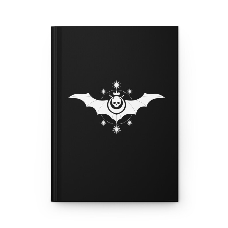 May include: Black notebook with a white bat and skull design. The bat has a crown on its head and is surrounded by a circle of stars.