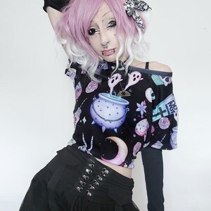 May include: Black off-the-shoulder crop top with a whimsical print featuring a cauldron, ghosts, and moons. The top is paired with a black skirt with a corset-style waist and a tulle overlay. The outfit has a gothic aesthetic.