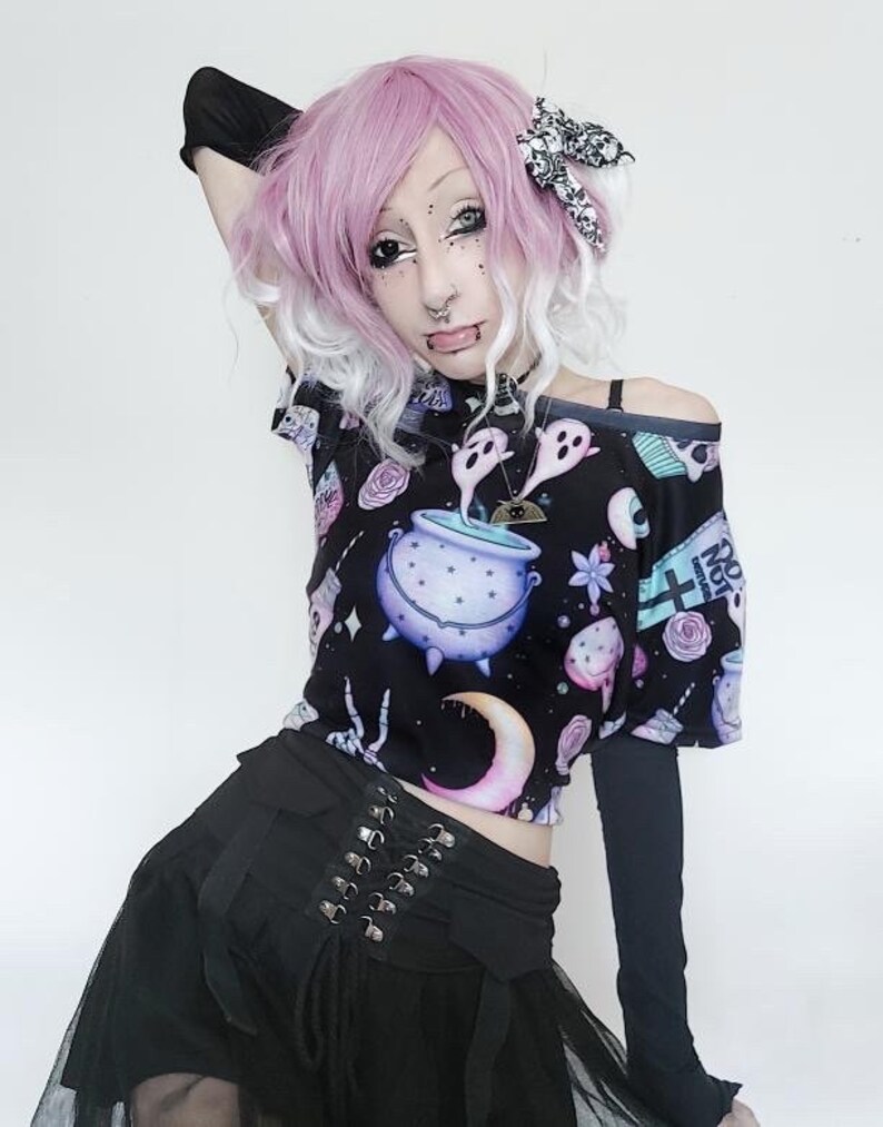 May include: Black off-the-shoulder crop top with a whimsical print featuring a cauldron, ghosts, and moons. The top is paired with a black skirt with a corset-style waist and a tulle overlay. The outfit has a gothic aesthetic.