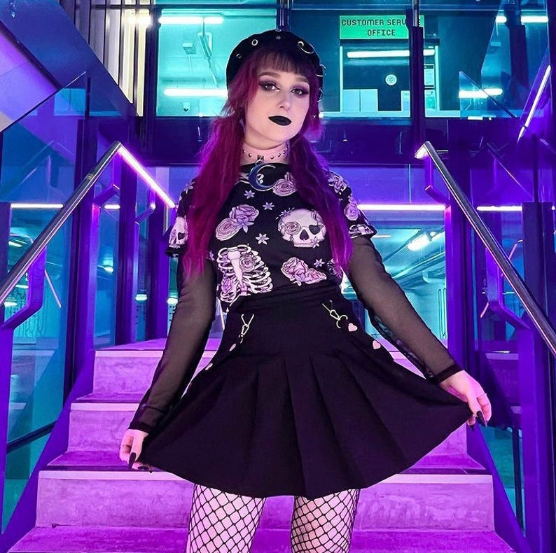 May include: A woman wearing a black and white skull and floral print crop top with a black mesh long-sleeved shirt, a black pleated skirt, and black fishnet tights. She is standing on a purple staircase with a sign that reads "Customer Service Office".