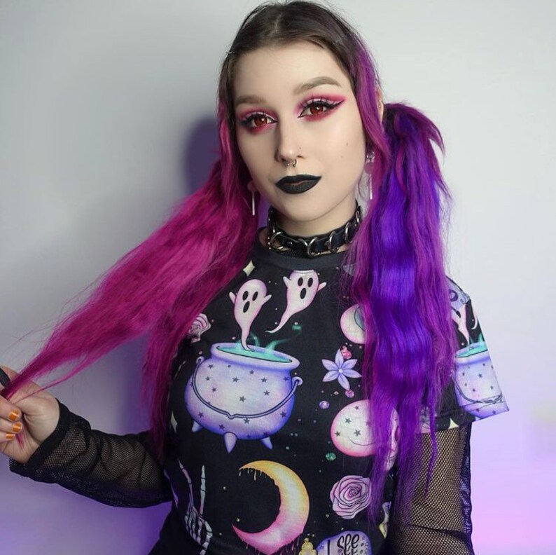 May include: A black t-shirt with a spooky cauldron and ghost design. The shirt features a round neck and short sleeves. The model is wearing a black choker and has purple and pink hair.
