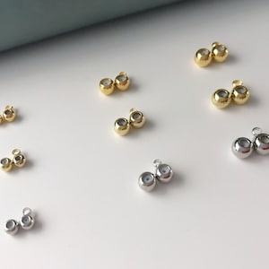 May include: Assortment of gold and silver bead caps with a hole in the centre. The bead caps are arranged in rows on a white surface.