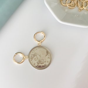 May include: A gold coin pendant with a portrait of a woman on one side and a design on the other side. The pendant is attached to a gold jump ring.
