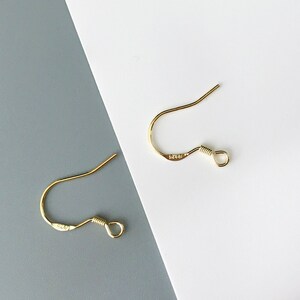 May include: Two gold-toned earring hooks with a small loop at the end. The hooks are on a white and gray background.