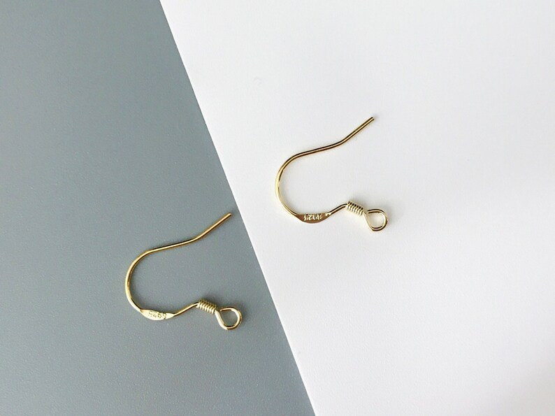 May include: Two gold-toned earring hooks with a small loop at the end. The hooks are on a white and gray background.