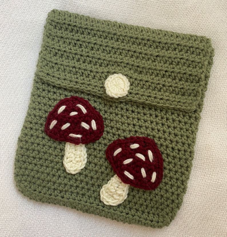 May include: A green crocheted pouch with a flap closure and a white button. The pouch features two embroidered mushroom appliques with red caps and white stems.