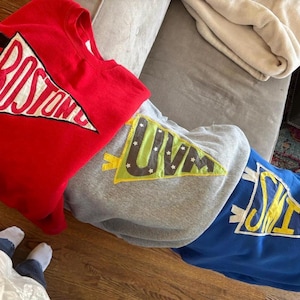 May include: Three sweatshirts in red, grey, and blue, each with a pennant design. The red sweatshirt displays "BOSTON" in white, the grey one says "UVM", and the blue one says "SMITH". The sweatshirts are folded on a grey surface.