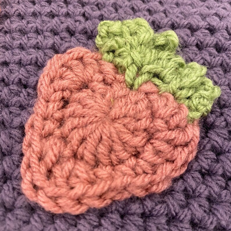 May include: A crocheted strawberry with a pink body and green leaves. The strawberry is made with yarn and has a textured surface.
