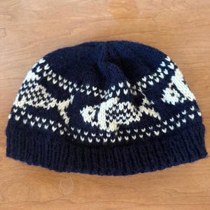 May include: A dark navy blue knit beanie featuring a repeating white pattern of stylized fish. The hat has a ribbed brim and a rounded crown, constructed with a thick yarn texture that displays a consistent, detailed knit stitch throughout.