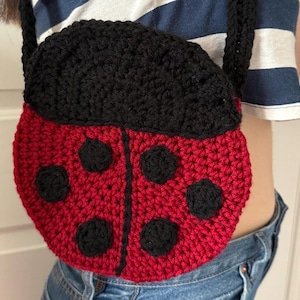 May include: A handmade crochet ladybug-shaped crossbody bag. The bag is primarily red with black accents, including the spots and flap. The strap is black. The bag is worn over the shoulder.