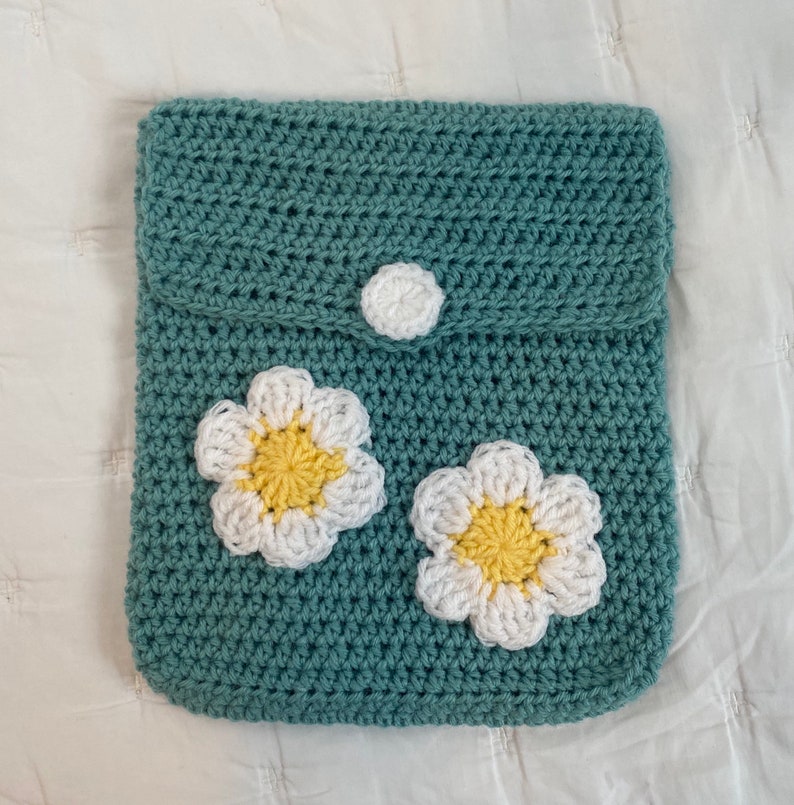 May include: A teal crocheted bag with a flap closure and a white button. The bag features two white crocheted flowers with yellow centers.