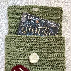 May include: A green crocheted book sleeve with a white crocheted flower and a red and white crocheted mushroom. The book sleeve is holding a book titled "The New York Times Bestseller The House".