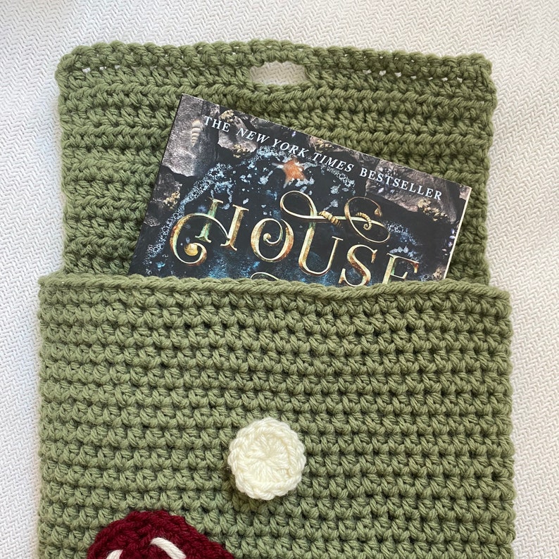 May include: A green crocheted book sleeve with a white crocheted flower and a red and white crocheted mushroom. The book sleeve is holding a book titled "The New York Times Bestseller The House".