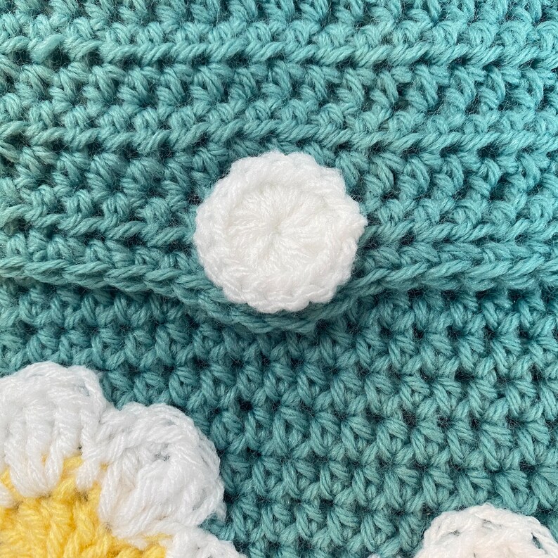 May include: Close-up of a teal crocheted bag with white and yellow crocheted flowers. The bag has a white crocheted button.