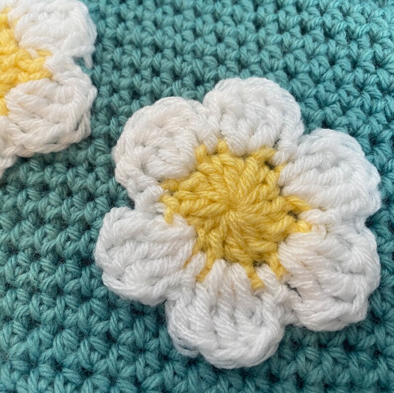 May include: Close-up of a crocheted flower with white petals and a yellow center. The flower is on a teal crocheted background.