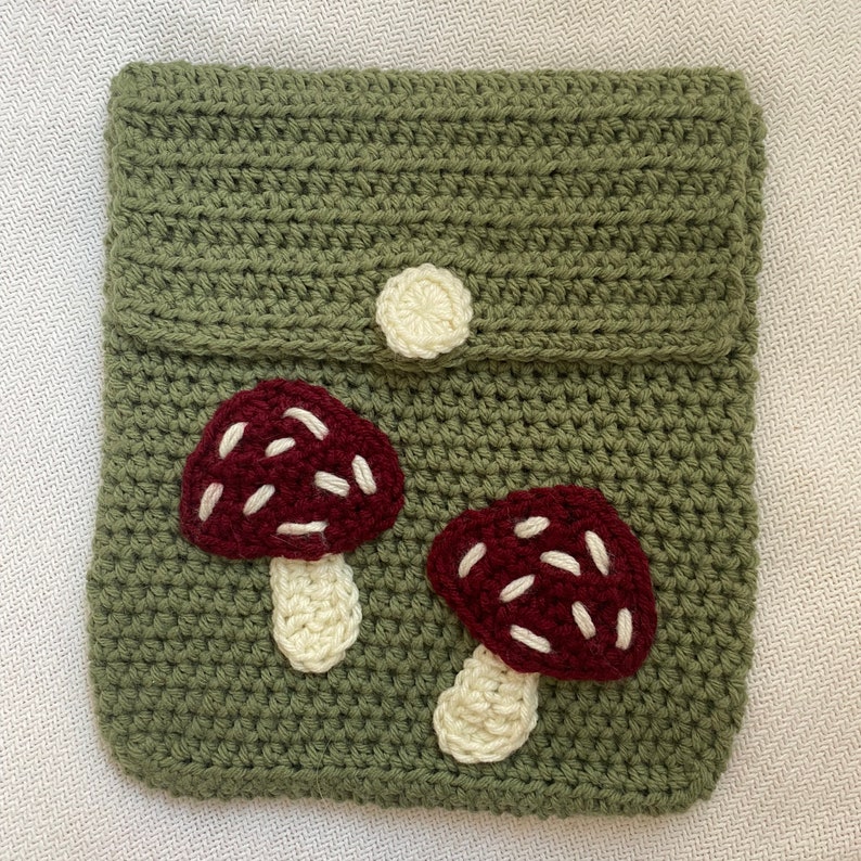 May include: A green crocheted pouch with a flap and a white button closure. The pouch features two embroidered red and white mushrooms.