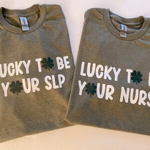 以下が含まれることがあります： 「Lucky to be your SLP」と「Lucky to be your Nurse」という白い文字が書かれた緑色のTシャツ2枚。各フレーズの横にクローバーのグラフィックが描かれている。