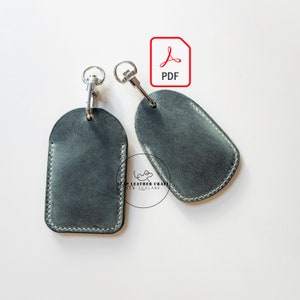 May include: Two dark gray leather keychains with silver key rings and a white stitch detail. The keychains are shaped like a tag and have a rounded top. The image also includes a red PDF icon.