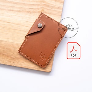 Pode incluir: Uma carteira de couro marrom com fecho de aba e um botão de metal. A carteira tem uma borda costurada e o nome da marca "Leap Leather Craft New Zealand" está estampado na frente.