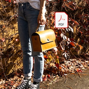 May include: A woman is holding a brown leather satchel bag with a flap closure and a metal buckle. She is wearing blue jeans and black high-top sneakers. The bag is sitting on a bed of fallen autumn leaves.