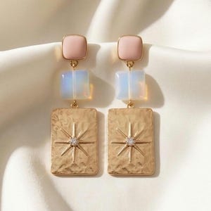 Puede incluir: Un par de pendientes dorados con diseño de estrella. Cada pendiente presenta una parte superior cuadrada rosa, un cuadrado translúcido y un colgante rectangular texturizado con una estrella y un pequeño cristal. Los pendientes se exhiben sobre una superficie de satén blanco.