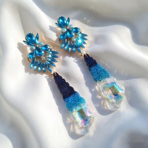 Può includere: Un paio di orecchini blu scintillanti. Ogni orecchino presenta un gruppo di cristalli blu e un design sfumato, che passa dal blu scuro al blu chiaro, con una base trasparente e iridescente. Sfondo bianco.