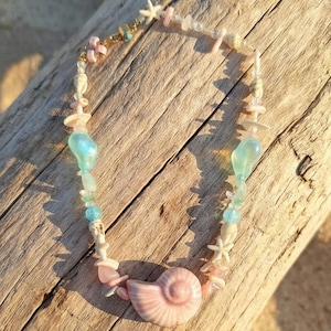 May include: A necklace featuring a large pink seashell pendant, complemented by small seashells, starfish, and beads in shades of pink, blue, and white. The necklace is displayed on a wooden surface, evoking a beachy theme.