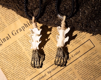 May include: Two necklaces with a skeletal hand pendant and a bone charm. The necklaces are made of black cord and have silver clasps. The pendants are made of metal and have a gold finish. The bone charms are white and have a natural finish.