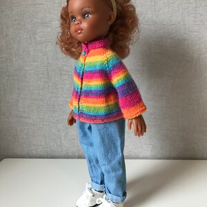 K&ouml;nnte beinhalten: Eine Puppe mit einem gestreiften Strickcardigan in Regenbogenfarben, hellblauen Jeans und wei&szlig;en Schuhen mit Punkten. Die Puppe hat braune Haut, lockiges rotes Haar und ein goldenes Stirnband. Der Cardigan hat einen Knopfverschluss.