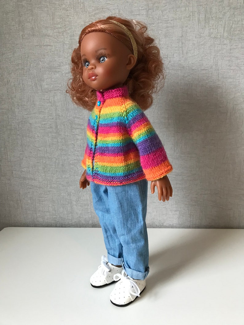 K&ouml;nnte beinhalten: Eine Puppe mit einem gestreiften Strickcardigan in Regenbogenfarben, hellblauen Jeans und wei&szlig;en Schuhen mit Punkten. Die Puppe hat braune Haut, lockiges rotes Haar und ein goldenes Stirnband. Der Cardigan hat einen Knopfverschluss.