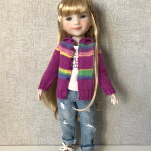 May include: A doll wearing a purple and rainbow striped cardigan, a white t-shirt with a graphic print, blue jeans with distressed details, and floral sneakers.
