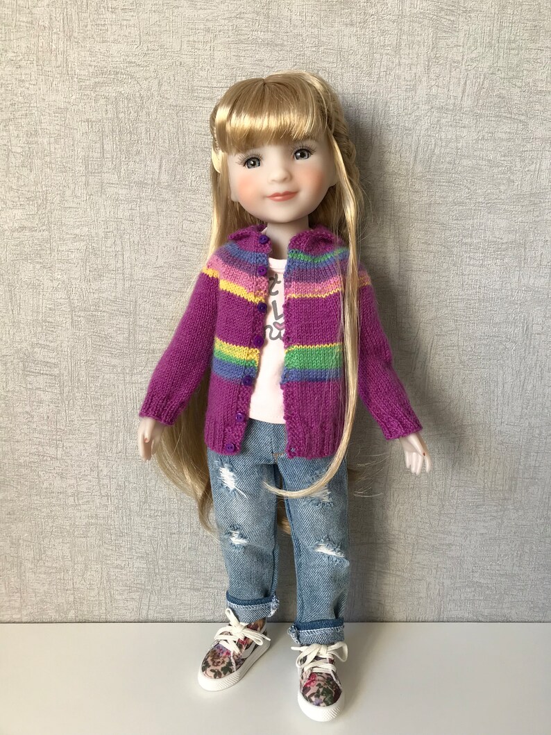 May include: A doll wearing a purple and rainbow striped cardigan, a white t-shirt with a graphic print, blue jeans with distressed details, and floral sneakers.