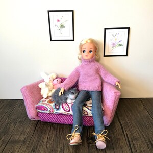 以下が含まれることがあります： ピンクのセーターを着て青いジーンズを履いたピンクのドールが、ピンクとパープルの模様のついたソファに座っています。ドールはブロンドの髪と青い目をしています。ソファにはぬいぐるみが3体置かれています。