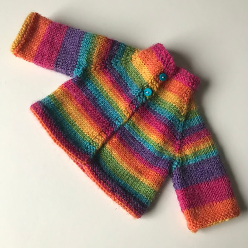 K&ouml;nnte beinhalten: Ein handgestrickter Baby-Cardigan in Regenbogenfarben mit roten, orangen, gelben, gr&uuml;nen, blauen und violetten Streifen. Der Cardigan hat eine Knopfleiste mit zwei t&uuml;rkisfarbenen Kn&ouml;pfen. Ein gem&uuml;tliches und fr&ouml;hliches Kleidungsst&uuml;ck.