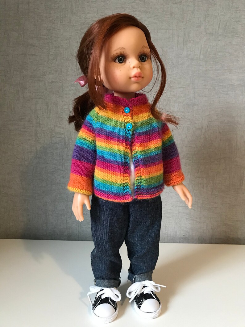 K&ouml;nnte beinhalten: Eine Puppe mit einem gestreiften Strick-Cardigan in Regenbogenfarben mit blauen Kn&ouml;pfen, dunkelblauen Jeans und schwarz-wei&szlig;en Turnschuhen. Die Puppe hat rotes Haar und einen Pferdeschwanz.