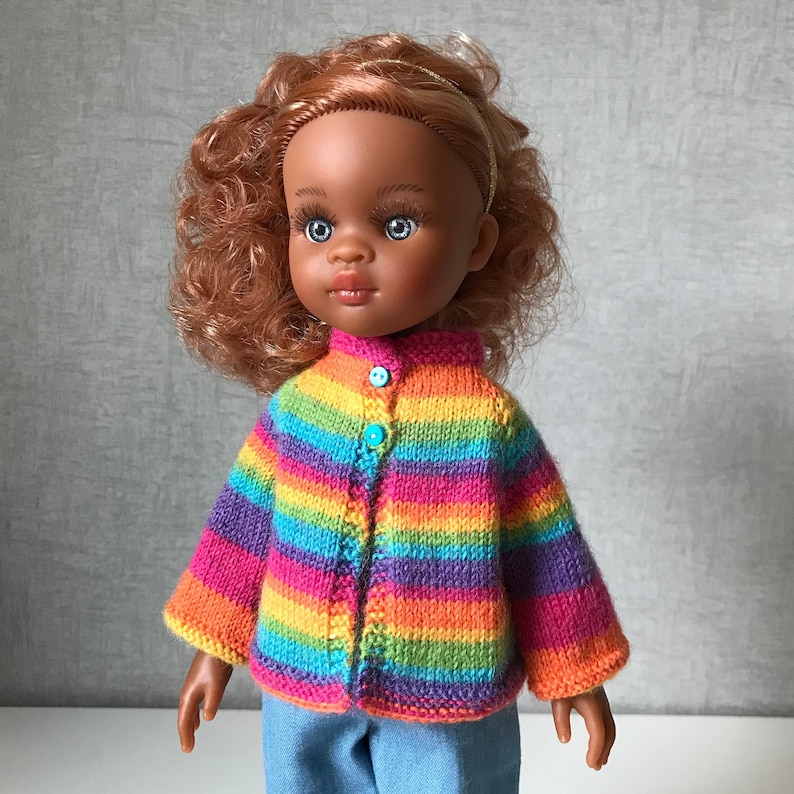 K&ouml;nnte beinhalten: Puppe mit brauner Haut, blauen Augen und roten Locken, die einen gestreiften Strickcardigan in Regenbogenfarben mit blauen Kn&ouml;pfen und Jeans tr&auml;gt. Der Cardigan hat Streifen in Pink, Orange, Gelb, Gr&uuml;n und Lila.