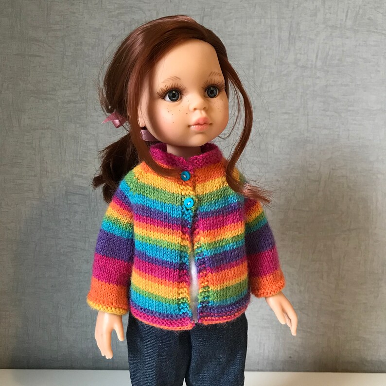 K&ouml;nnte beinhalten: Eine Puppe tr&auml;gt eine gestreifte Strickjacke in Regenbogenfarben mit drei blauen Kn&ouml;pfen. Die Puppe hat rotes Haar, Sommersprossen und tr&auml;gt eine blaue Jeans. Die Strickjacke hat horizontale Streifen in verschiedenen Farben.