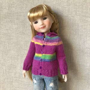 May include: A doll wearing a purple, green and yellow striped cardigan with buttons down the front. The cardigan is knitted and has a soft, fuzzy texture.