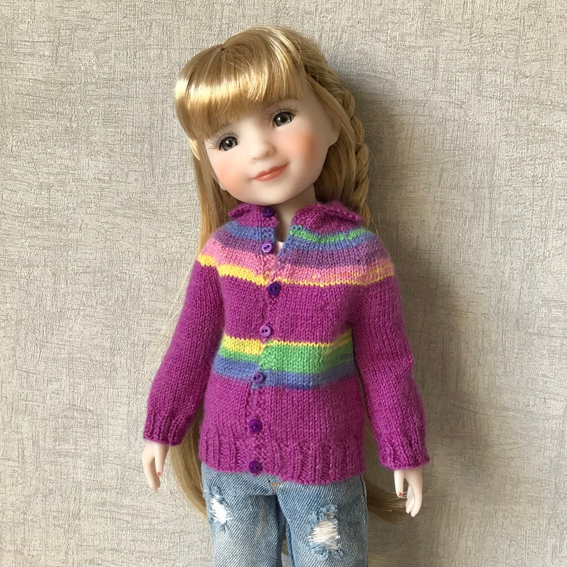May include: A doll wearing a purple, green and yellow striped cardigan with buttons down the front. The cardigan is knitted and has a soft, fuzzy texture.