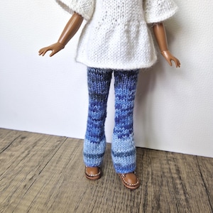 May include: A doll wearing a white knit top and blue knit pants. The pants have a gradient of blue hues. The doll is also wearing brown leather shoes with white stitching. The outfit is handmade.