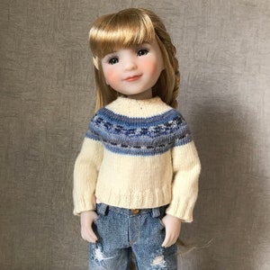 Könnte beinhalten: Eine Puppe trägt einen cremefarbenen Pullover mit einem blauen und grauen Musterband um den Halsausschnitt. Die Puppe hat blonde Haare und trägt eine blaue Jeans.