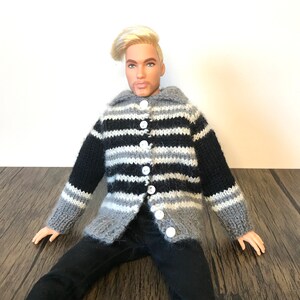 May include: A doll wearing a button-up, striped sweater in black, gray, and white. The sweater has a collar and is buttoned up with white buttons. The doll is also wearing black pants. The doll has blonde hair and is sitting on a wooden surface.