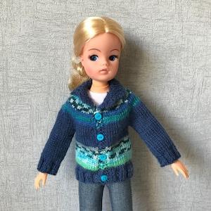 Puede incluir: Un cárdigan azul de punto con un estampado de rayas de colores y botones azules. El cárdigan lo lleva una muñeca con el pelo rubio.