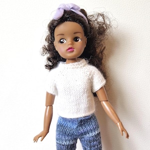 May include: A doll with dark skin, brown curly hair, and a lavender headband. The doll wears a white knit t-shirt and blue knit pants. The doll's arms are articulated.