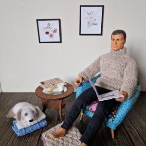 May include: A man wearing a beige sweater sits in a blue armchair with floral fabric. He is reading a newspaper and holding a cup of coffee. A small table with a plate of cookies and a cup of coffee sits next to the chair. There are two framed pictures on the wall behind the man.